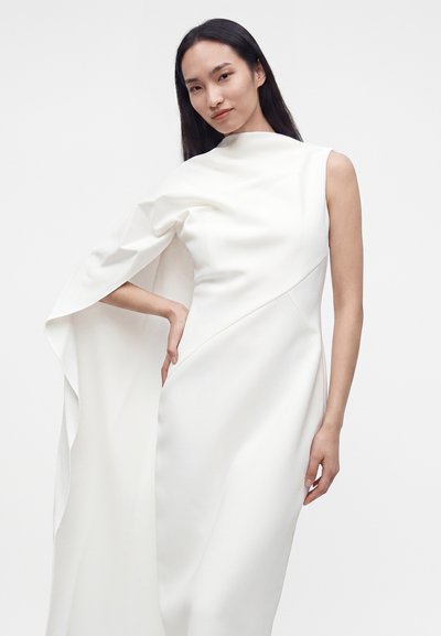 Femme en robe blanche sans manches avec une manche fluide en forme de cape sur un côté, debout, main sur la hanche, devant un fond uni.