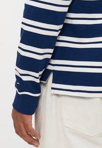 Marinedempur og hvite striper langermet skjorte med sideslitt iført off-white bukser, viser venstre arm og hånd.