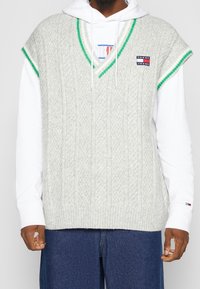 Graue, kabelgestrickte Weste mit V-Ausschnitt und grünem Rand, getragen über einem weißen Hoodie. Verfügt über ein kleines Marken-Patch auf der linken Brust.