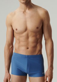 Niebieskie szorty kąpielowe o gładkiej teksturze, dopasowanym kroju i krótkim kroju nogawki. Zawierają subtelne logo z boku jako element brandingu.