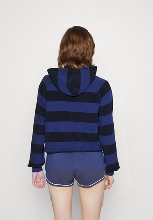 Person standing facing away wearing a blue and black striped hoodie and dark blue athletic shorts with white trim.