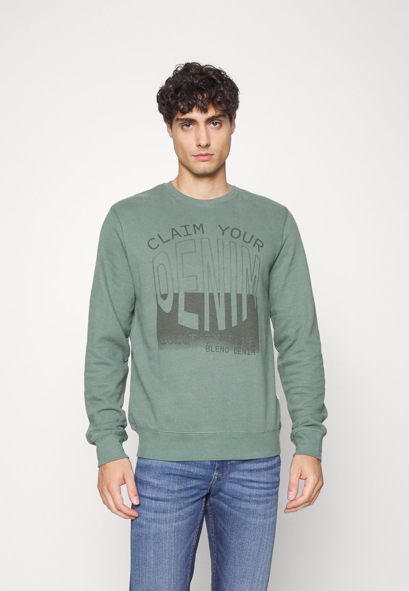 Sudadera de algodón verde con cuello redondo y mangas largas. Presenta una gran impresión gráfica negra que dice "RECLAMA TU DENIM."