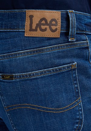 Vedere din spate a blugilor albaștri din denim cu patch maro deschis din piele și o mică etichetă de material, ambele afișând numele brandului Lee deasupra buzunarului din spate.