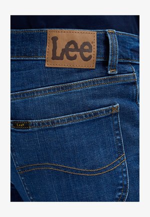 Vedere din spate a blugilor albaștri din denim cu patch maro deschis din piele și o mică etichetă de material, ambele afișând numele brandului Lee deasupra buzunarului din spate.