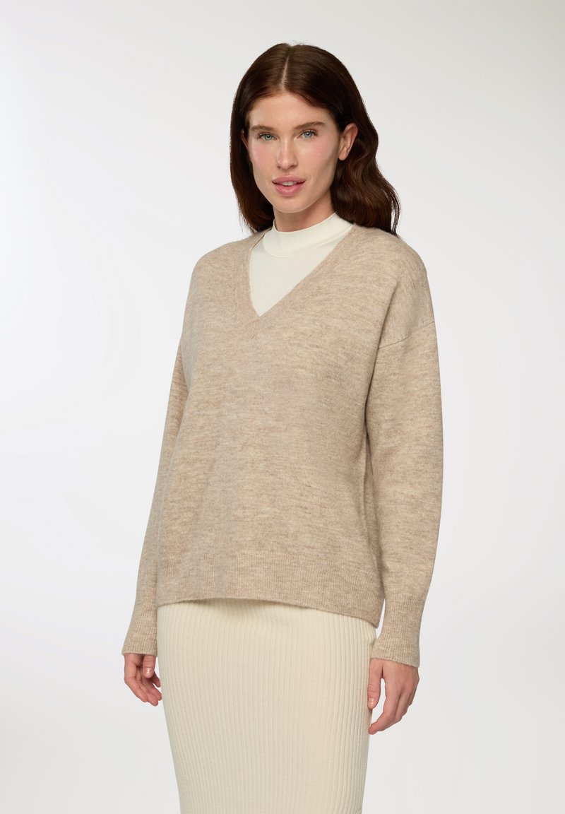 Beżowy sweter w serek wykonany z miękkiego materiału, z obniżonymi ramionami, długimi rękawami i luźnym krojem, zestawiony z kremową spódnicą w prążki.