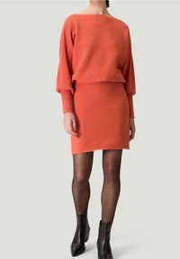Femme portant une robe côtelée de couleur corail orange avec des manches longues, des collants noirs transparents et des bottines noires, se tenant sur un fond neutre.