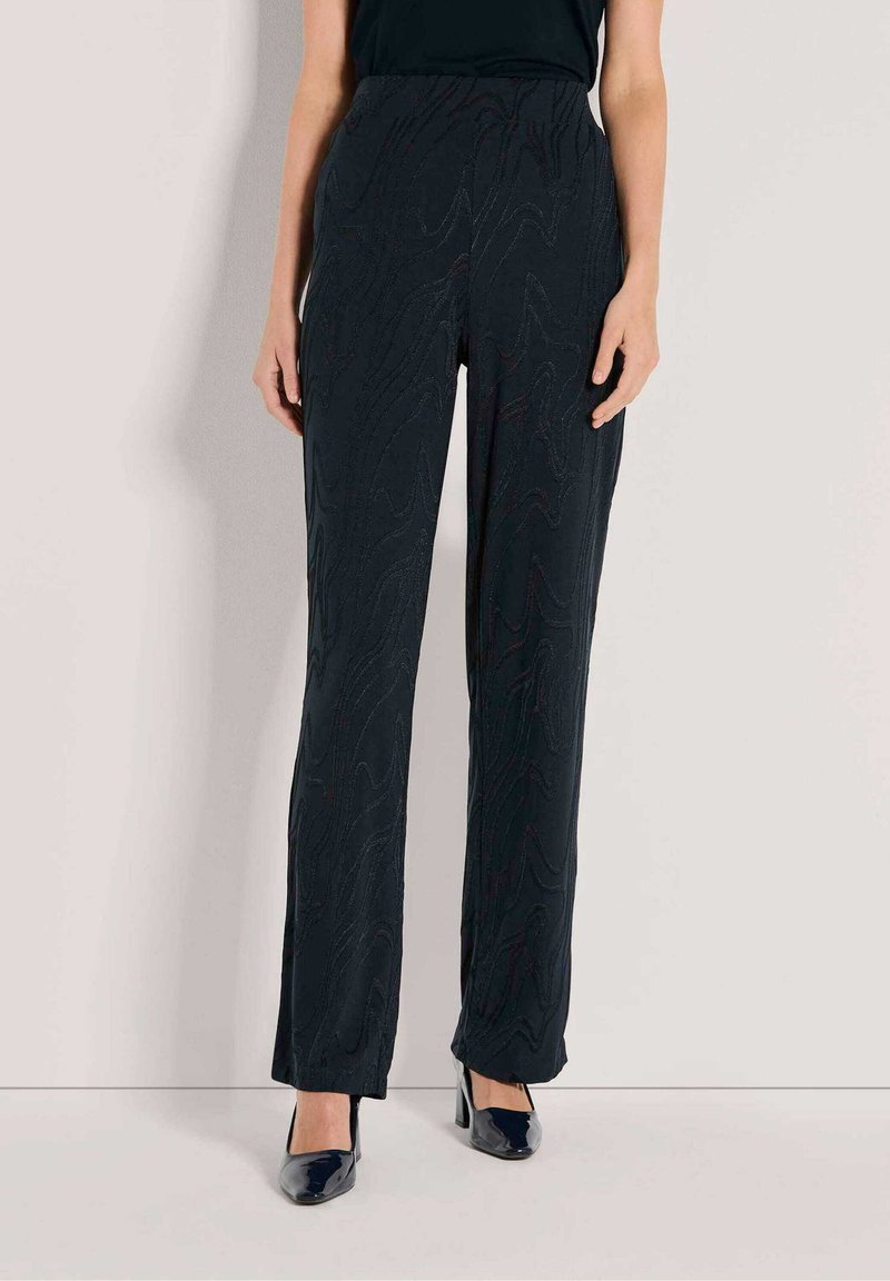 Vrouw in zwarte gestructureerde rechte broek en zwarte hoge hakken, staand tegen een effen lichte achtergrond.