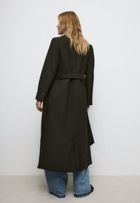Cappotto lungo di un verde scuro con vita cintata, vestibilità su misura e maniche ampie. Abbinato a jeans chiari a campana con orli sfilacciati.