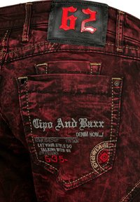 Rode spijkerbroek van denim met een verouderde textuur, voorzien van gele stiksels, een zwart leren label met "62" en geborduurde merkinformatie op de achterzak.