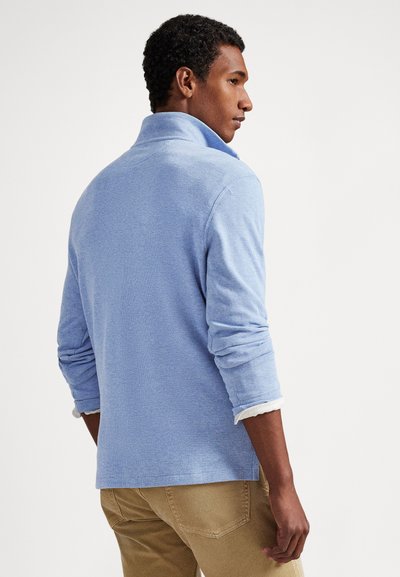 Vīrietis, skatoties sānis, valkā gaiši zilu džemperi ar garām piedurknēm virs baltas krekla ar sarullētām piedurknēm un bēšas bikses uz vienkārša balta fona.