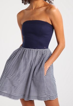 Trägerloses dunkelblaues Kleid mit tailliertem Oberteil und ausgestelltem Rock mit horizontalen weißen und marineblauen Streifen, Hand in der Tasche.