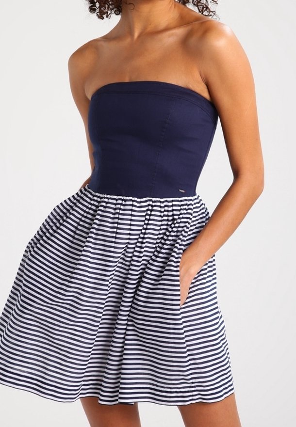 Robe bleu marine sans bretelles avec un corsage ajusté et une jupe évasée comportant des rayures horizontales blanches et bleu marine, main dans la poche.