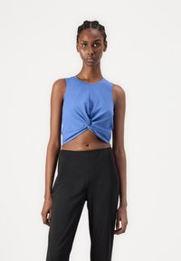 Sinine lühike tank top, mille esiosas on keeratud sõlm, koos kõrge vöökohaga mustade pükstega. Sile puuvillane materjal.