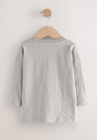 Jasnoszara koszula z długim rękawem o okrągłym dekolcie, z gładką fakturą i subtelnym przeszyciem, zawieszona na drewnianym wieszaku.