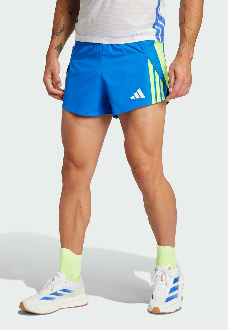 Modré sportovní šortky s žlutými akcenty a bílým logem Adidas, s hladkým textilním povrchem a bočními rozparky pro lepší pohyblivost, nošené s bílými botami a neonovými ponožkami.
