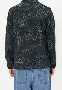 Osoba w ciemnej polarowej kurtce z abstrakcyjnymi zielonymi i szarymi wzorami, stojąca tyłem do kamery, w połączeniu z jasnoniebieskimi dżinsami.