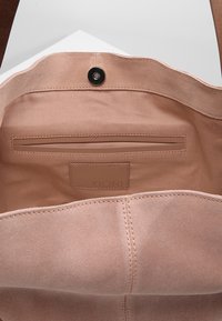 Ljusrosa mocka toteväska med en slät inredning, som har en ficka med dragkedja och en varumärkesetikett. Magnetknäppning högst upp.