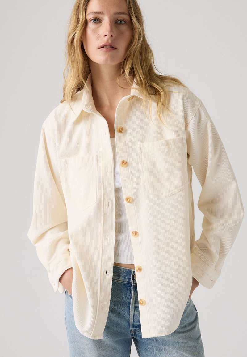 Femme portant une chemise boutonnée en velours côtelé écrue sur un haut blanc et un jean bleu clair, debout devant un fond clair uni.