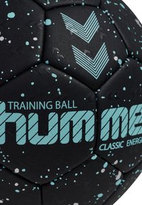 Czarna piłka treningowa z turkusowym i białym wzorem rozprysków, wyposażona w teksturowane panele, wyraźne oznaczenia i graficzne strzałki kierunkowe.