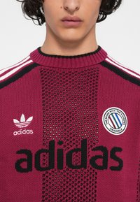 Tuvo plāns uz cilvēka, kurš valkā bordo Adidas džemperi ar melnu acs rakstu, balto logo, zīmola uzšuvi un trim baltām svītrām uz pleciem.