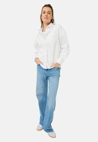 Camicia bianca con bottoni e tasca, abbinata a jeans svasati blu chiaro e sneakers bianche, esposta su uno sfondo neutro.