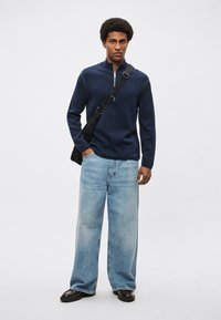 Tengerészkék, cipzáras kötött pulóver finom varrással, világoskék, bőszárú farmerral és sötét cipővel párosítva. Állítható fekete oldaltáska.