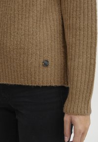 Brown ribbed knit sweater with a cropped hem and a black button accent on the side. Soft texture, long sleeves, and close-fitting silhouette.