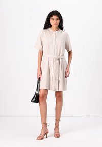 Beige linen shirt dress with short sleeves, button front, and waist tie detail. Paired with a black handbag and strappy tan heels.