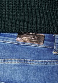 Pull en maille côtelée vert foncé reposant sur un jean en denim bleu clair avec une étiquette en cuir marron "ONLY" sur la taille.