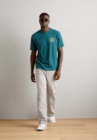 Teal bomulds t-shirt med grafisk design på fronten. Båret med lysegrå jeans og hvide sneakers. Modellen bærer mørke solbriller.