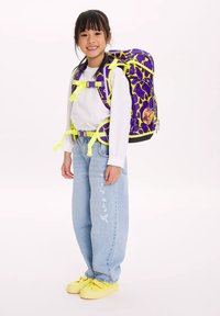 Ragazza sorridente che indossa una maglietta bianca, jeans azzurri chiari, scarpe gialle e uno zaino grande viola con cinghie gialle e motivi.
