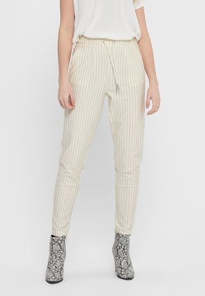 Vrouw draagt crèmekleurige, slimfit broek met dunne verticale strepen en enkellaarzen met slangenhuidpatroon, staand tegen een effen achtergrond.