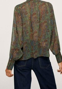 Blouse légère à motif paisley vert, coupe ample, manches longues avec poignets boutonnés. Associée à un jean bleu foncé avec poches arrière.