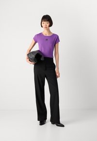 Purple fitted t-shirt with a small black logo, paired with high-waisted black trousers. Model holds a black clutch. Smooth fabrics, contemporary fit.
