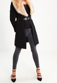 Sort frakke med faux fur krave, bælte i taljen, lagdelt over en sort blondetop. Grå skinny jeans og sorte strop højhælede sko fuldender outfittet.