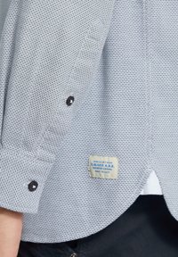 Chemise texturée bleu clair avec un poignet boutonné et une étiquette blanche comportant des informations sur la marque cousue en bas de l'ourlet.