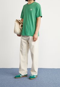 Grön bomullströja med en liten vit logga, beige löst sittande byxor och gröna skor med vita ränder. Beigefärgad tote-väska hålls i handen.