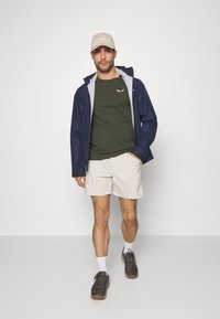 Marine vanntett jakke med grå foring, olivengrønn t-skjorte, beige shorts og brune tursko. Slim-fit design og avslappet utendørs stil.