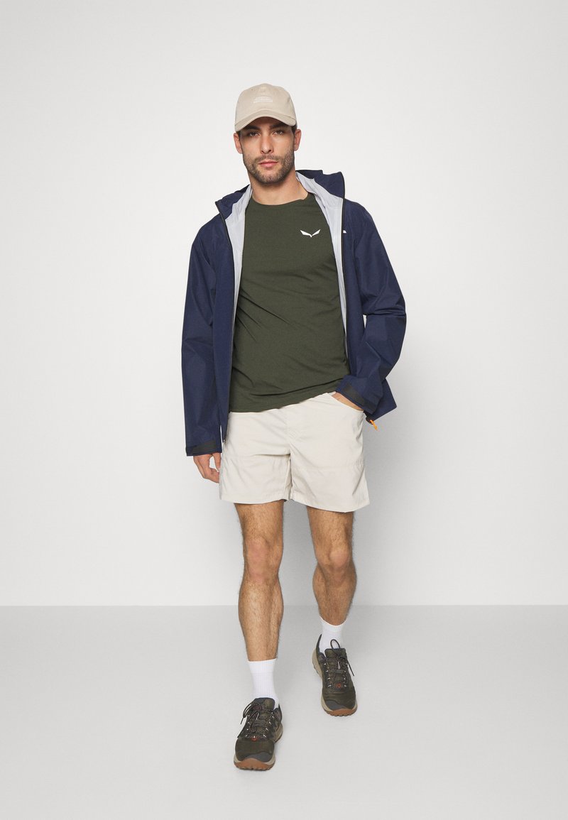 Marine vanntett jakke med grå foring, olivengrønn t-skjorte, beige shorts og brune tursko. Slim-fit design og avslappet utendørs stil.