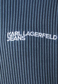 Țesătură în dungi de un albastru închis, cu nuanțe variate. Prezintă text brodat alb "KARL LAGERFELD JEANS." Textura este ribbată și țesută.
