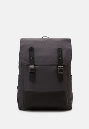 Sac à dos gris foncé avec rabat supérieur, deux sangles noires à boucles et une poche avant avec panneau de base noir, présenté sur un fond clair.