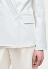 Gros plan d'un blazer blanc avec un bouton et une poche à rabat, porté sur un pantalon blanc assorti, montrant une main détendue sur le côté.
