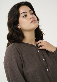 Hnědá, texturovaná blůza s kulatým výstřihem, bílými knoflíky a nařasenými manžetami. Modelka lehce sahá na límec.