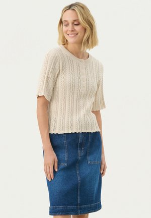 Beige kötött pulóver rövid ujjal, bordázott mintázattal és öt gombbal elöl; kék farmer szoknyával párosítva.
