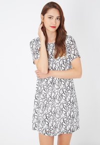 Mujer joven con cabello largo castaño, vistiendo un vestido blanco de mangas cortas con patrones circulares abstractos en negro, de pie contra un fondo blanco.