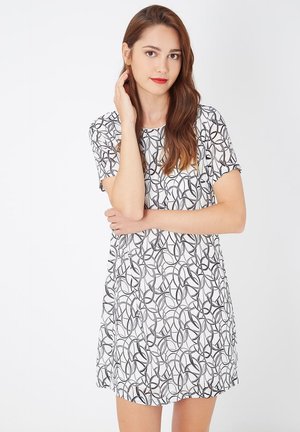 Mujer joven con cabello largo castaño, vistiendo un vestido blanco de mangas cortas con patrones circulares abstractos en negro, de pie contra un fondo blanco.