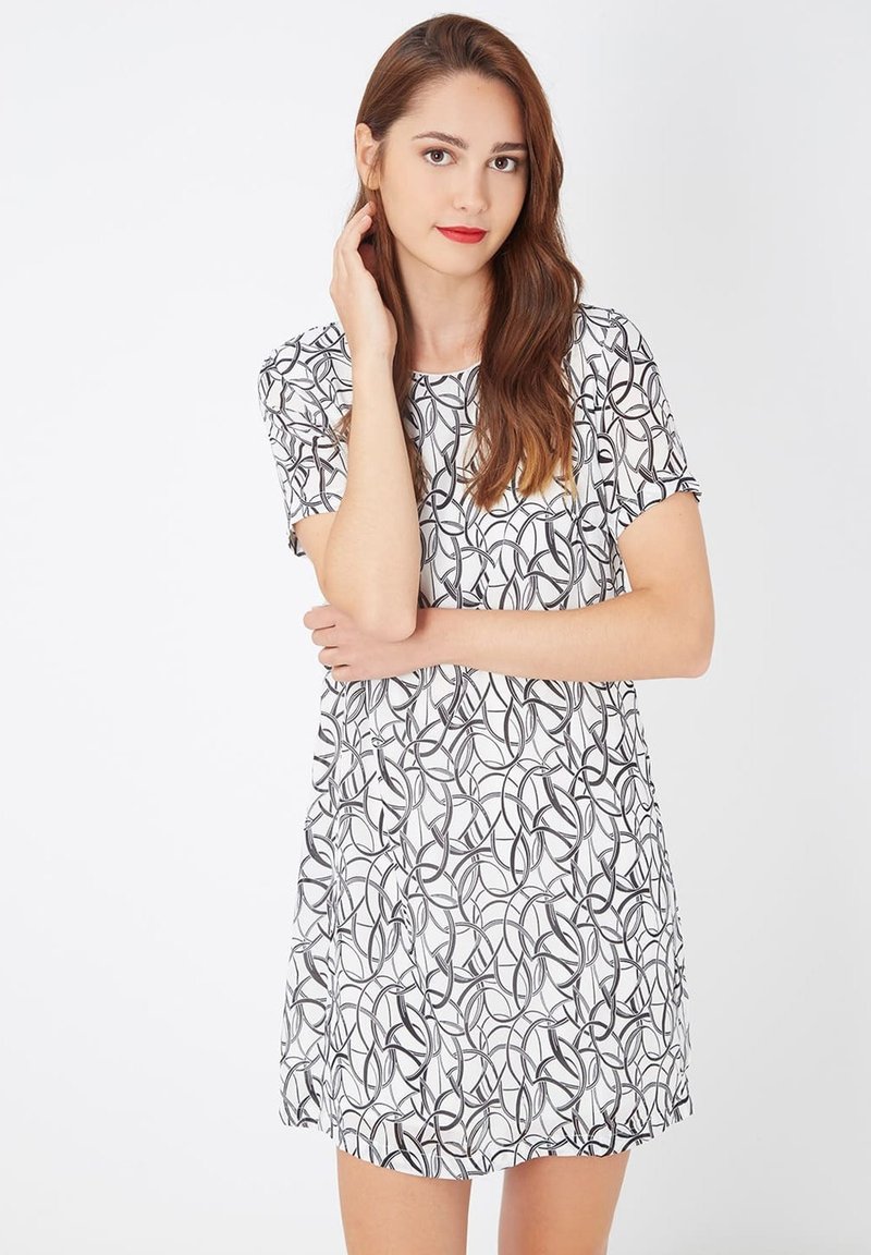 Mujer joven con cabello largo castaño, vistiendo un vestido blanco de mangas cortas con patrones circulares abstractos en negro, de pie contra un fondo blanco.