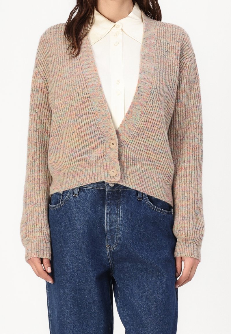 Personne portant une chemise crème boutonnée sous un cardigan en tricot beige tacheté avec des boutons en bois et un jean bleu taille haute.