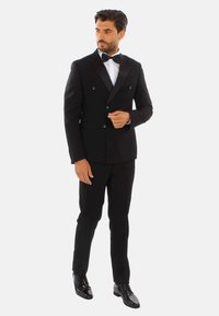 Giacca da smoking nera doppiopetto con rever a scialle, abbinata a pantaloni coordinati e scarpe eleganti nere lucide. Camicia bianca e cravatta a farfalla.