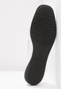 Baskets blanches avec une semelle en caoutchouc noir texturé présentant des motifs circulaires et un logo Geox. La partie supérieure est lisse avec un design minimaliste.